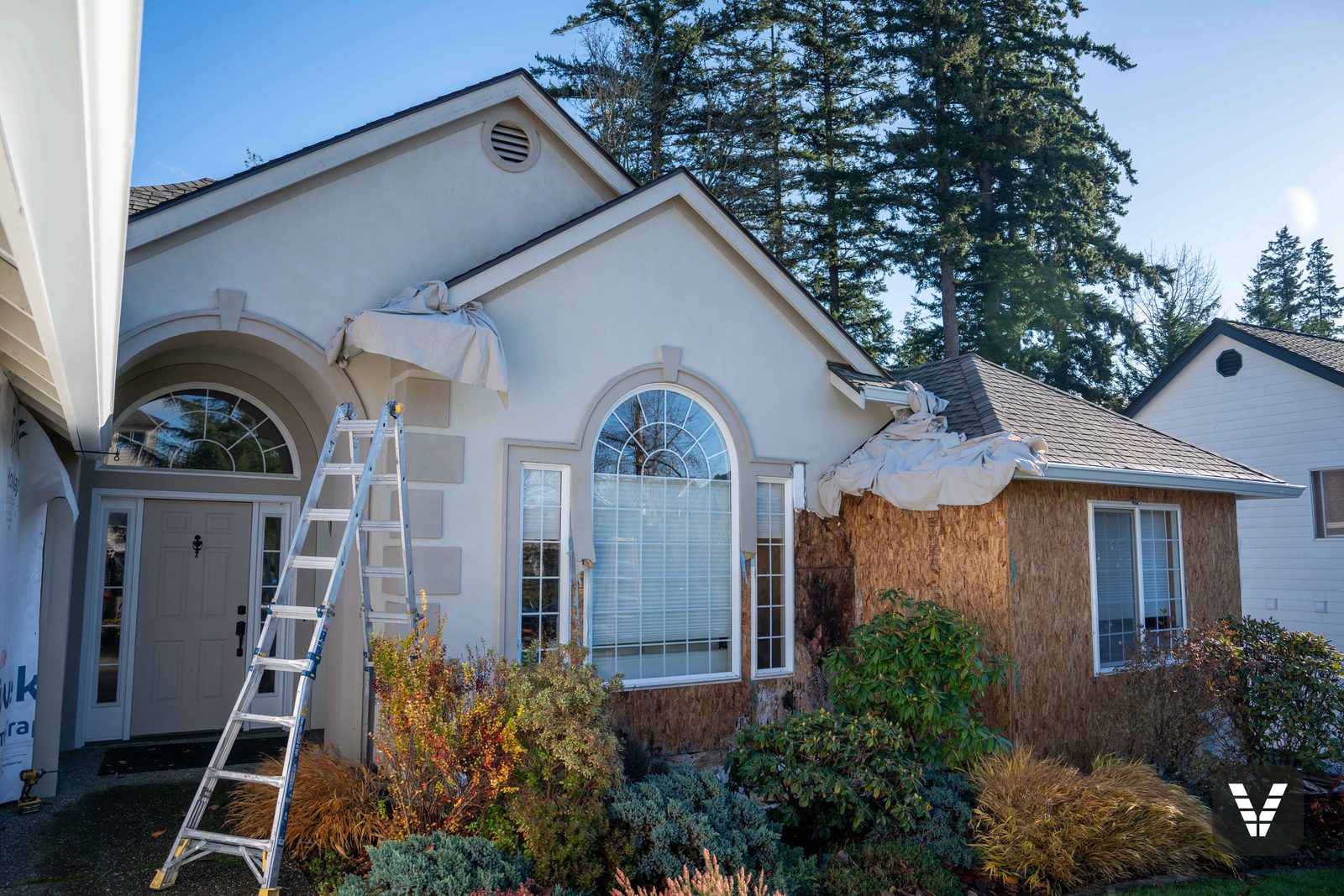 Tear-off in progress at the entry — stucco stripped, sheathing exposed, drop cloths protecting the garden beds below